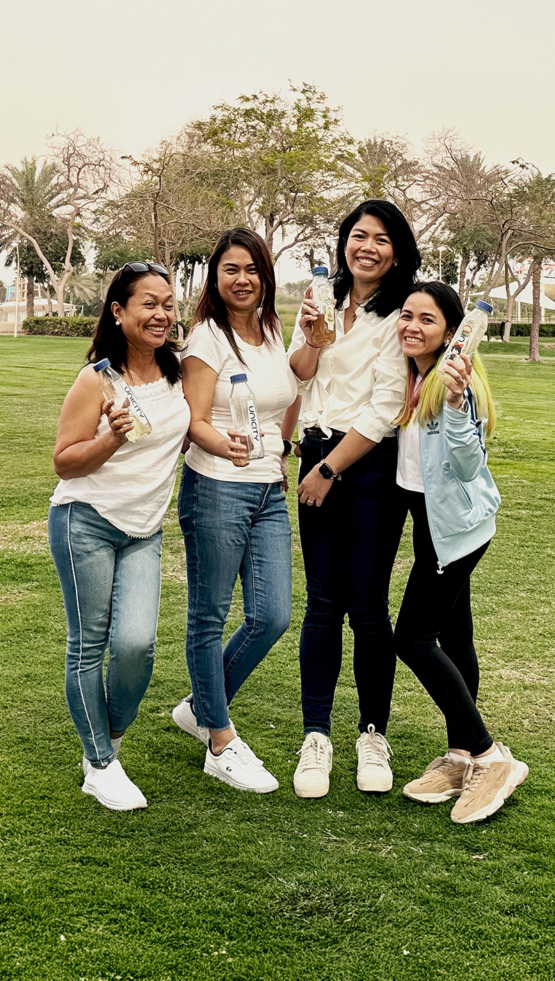 Four women walking together across a park lawn, laughing and holding drinks